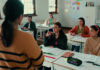 Why curriculum developments in ELT often fail A classroom scene with a teacher standing in front of a group of students sitting at desks, listening attentively. The students are diverse and appear engaged, with books and papers on their desks. The background shows educational posters on the walls and cabinets in the room.