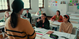 A classroom scene with a teacher standing in front of a group of students sitting at desks, listening attentively. The students are diverse and appear engaged, with books and papers on their desks. The background shows educational posters on the walls and cabinets in the room.