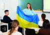Teaching displaced learners A teacher holding the Ukrainian flag stands in front of a classroom of students seated at desks, attentively listening to her lecture.