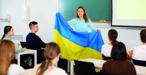 A teacher holding the Ukrainian flag stands in front of a classroom of students seated at desks, attentively listening to her lecture.