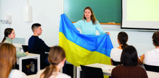 A teacher holding the Ukrainian flag stands in front of a classroom of students seated at desks, attentively listening to her lecture.