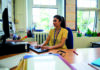 Narrative lesson planning A woman wearing a yellow dress and a lanyard sits at a desk, working on a computer in an office with large windows and various office supplies and papers around her.