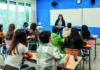 Is ‘research’ a dirty word? A teacher standing at the front of a classroom, smiling as she speaks to a group of seated students. The room has blue walls, a large window with natural light, and a whiteboard behind the teacher. The students are sitting at desks with notebooks and laptops, attentively listening.
