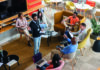 What do school leaders need to know about culture? A diverse group of people sitting in a modern, spacious lounge area are having a casual meeting. One person is standing and speaking to the others, who are seated in a circle with notebooks and electronic devices. The room is decorated with plants, and the seating consists of comfortable chairs and sofas around a central low table with glasses of water and papers.
