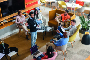 A diverse group of people sitting in a modern, spacious lounge area are having a casual meeting. One person is standing and speaking to the others, who are seated in a circle with notebooks and electronic devices. The room is decorated with plants, and the seating consists of comfortable chairs and sofas around a central low table with glasses of water and papers.