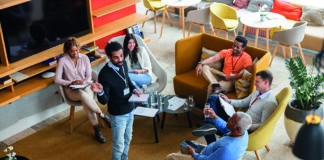 A diverse group of people sitting in a modern, spacious lounge area are having a casual meeting. One person is standing and speaking to the others, who are seated in a circle with notebooks and electronic devices. The room is decorated with plants, and the seating consists of comfortable chairs and sofas around a central low table with glasses of water and papers.
