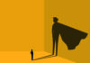 Small but mighty A small figure stands confidently with arms crossed, casting a large shadow on the wall resembling a superhero with a cape, in a minimalist style with a warm yellow background.