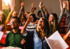 The seven superpowers of successful students A group of diverse students in a classroom cheering with excitement, their arms raised in celebration, while holding notebooks and papers.