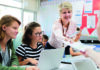 Uncovering a hidden world A teacher hands a worksheet to a student across a classroom table, where other students are seated with laptops in front of them. The setting is bright and colorful with educational materials on the walls.