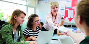 A teacher hands a worksheet to a student across a classroom table, where other students are seated with laptops in front of them. The setting is bright and colorful with educational materials on the walls.