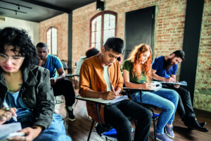 A diverse group of students seated in a classroom with brick walls, concentrating on writing in notebooks at their desks.