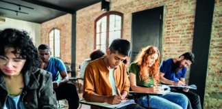 A diverse group of students seated in a classroom with brick walls, concentrating on writing in notebooks at their desks.