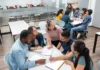 Collaborative conversations A group of students sitting around a table in a classroom, discussing with a teacher who is leaning over to assist them. Other groups of students in the background are similarly engaged in discussions, creating a collaborative learning environment.