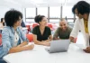 Don’t let it get away! A group of four people sitting around a table in a classroom setting, engaged in a discussion while looking at a laptop screen; two women are writing notes while the other two people are standing, one pointing at a document.