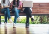 Going multimodal Three people sitting on a wooden bench, working on electronic devices; two are using tablets while the third is using a laptop. They are casually dressed in jeans and sneakers, with a leafy background visible.