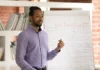 Rethinking the count-mass distinction beyond the canon of ELT grammar A person wearing a light purple shirt stands beside a flip chart displaying English grammar tenses, speaking to an audience in a room with bookshelves and a brick wall.