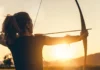 Utilising other strings in our bow A person practicing archery, drawing a bow with an arrow aimed forward against a sunset backdrop, highlighting the silhouette and arm guard.