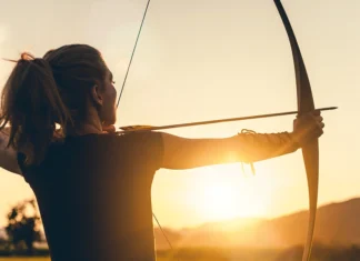 Utilising other strings in our bow A person practicing archery, drawing a bow with an arrow aimed forward against a sunset backdrop, highlighting the silhouette and arm guard.