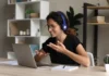 Teaching beyond borders A woman with headphones sits at a desk in front of a laptop, gesturing while engaged in a video call. She is smiling and appears to be in a modern home office with shelves in the background.