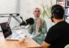 Voicing urban treasures Two people wearing headphones are seated at a microphone for a podcast recording; one, a woman in a hijab, speaks animatedly, while the man listens.