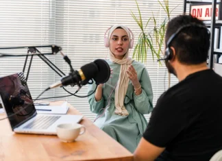 Voicing urban treasures Two people wearing headphones are seated at a microphone for a podcast recording; one, a woman in a hijab, speaks animatedly, while the man listens.