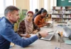 Culturally responsive leaders A group of people sitting at a table in an office setting, using laptops. One man is pointing at a screen while discussing something with a woman wearing a headscarf who is taking notes. In the background, another woman is focused on her laptop, with bookshelves visible behind her.