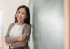 I am more than my Filipino accent A smiling woman stands in front of a chalkboard, holding a piece of chalk, wearing a beige cardigan over a white top and jeans, with long black hair.