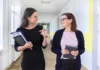 Find your people Two women walking and talking in a hallway; one is holding a clipboard and the other a notebook.