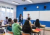 Holidays in a box A classroom with students seated at desks and a teacher standing near a whiteboard, clapping hands with a student next to them, as other students applauding; laptops and notebooks are visible on the desks.