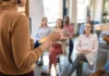 Listening our way forward A woman in a brown sweater holding a pen is speaking to a group of seated people in a classroom or seminar setting, with a focus on several attendees listening attentively.