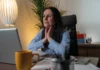 Our voice A woman sitting at a desk with a laptop, holding her neck and appearing to be in discomfort, with a yellow mug, lamp, and office items in the surroundings.