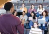 What does methodology mean to you? A teacher standing in front of a group of students in a lecture hall, gesturing as they appear to be in discussion or giving a lecture. The students are seated with laptops and notebooks, attentively listening.