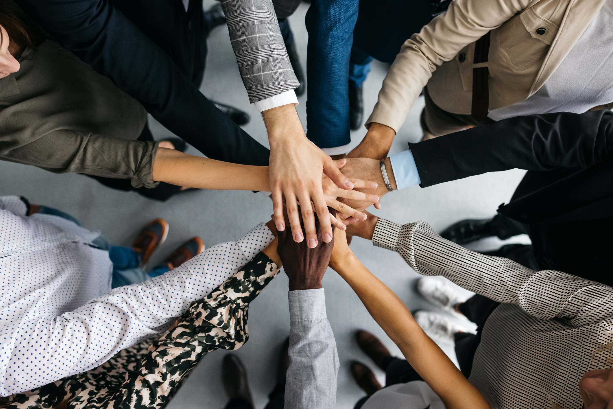 A group of people standing in a circle, each reaching in to place their hand on top of each other in the center, symbolizing unity and teamwork.