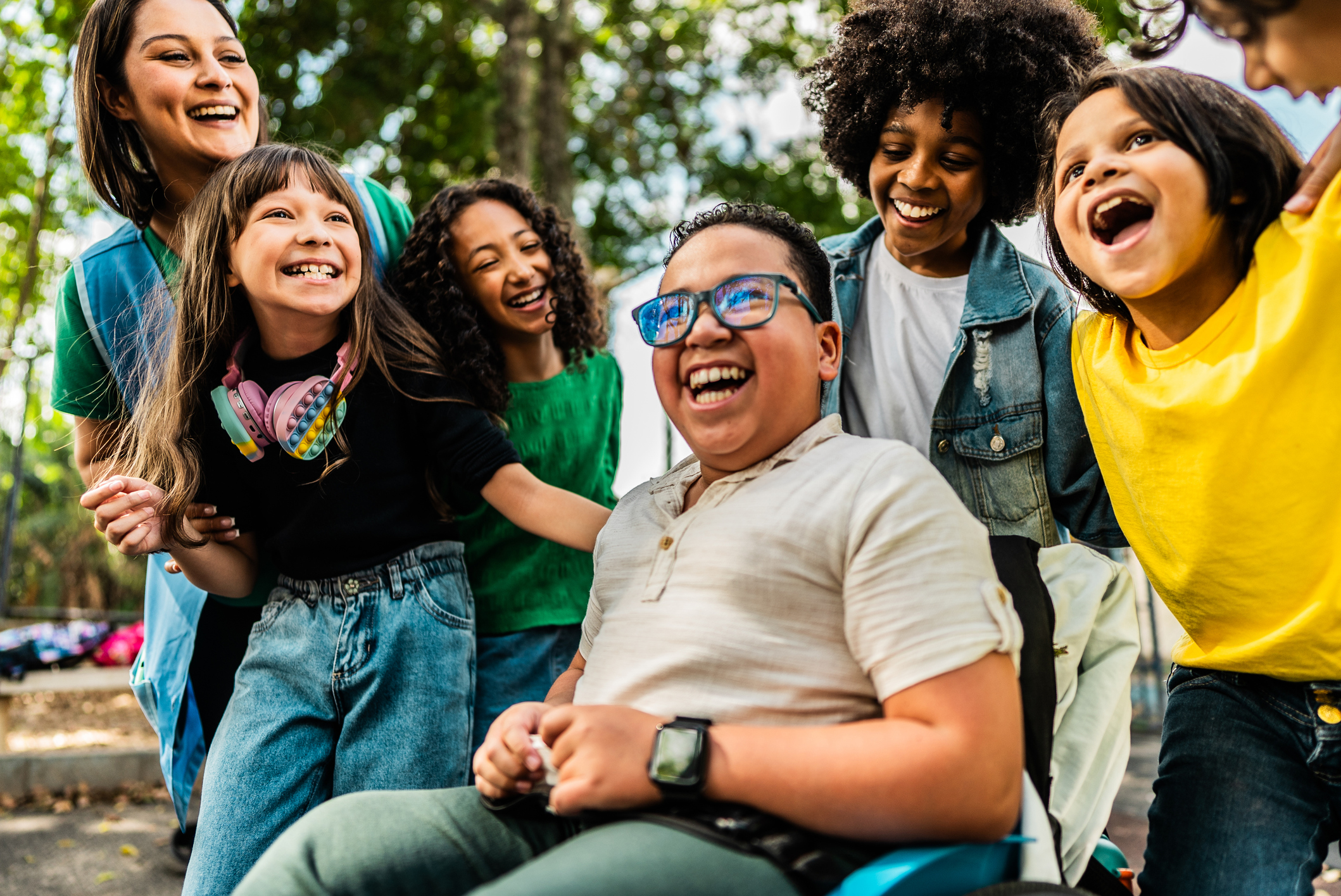 A group of diverse children and a woman are laughing and smiling together outside, with a child in a wheelchair at the center of the group.