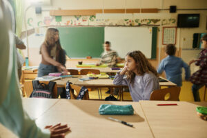A classroom with several children, one of whom is sitting at a desk looking away thoughtfully, while others move around energetically. A green chalkboard and colourful wall decorations are in the background.