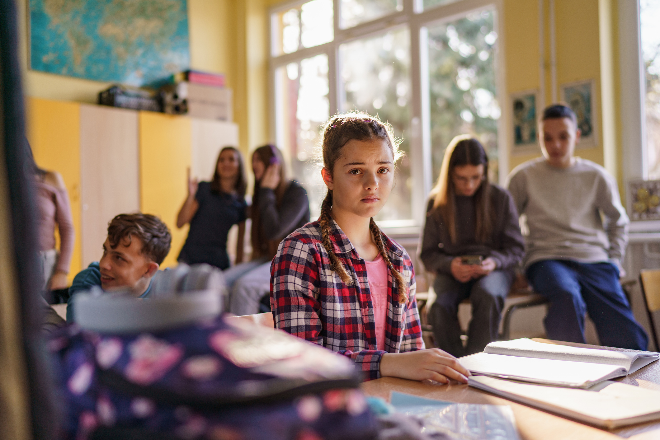 A young girl with braided hair and a plaid shirt sits at a desk in a classroom, looking towards the camera with a serious expression. Behind her, several students are sitting on desks, some looking at their phones while others are talking.