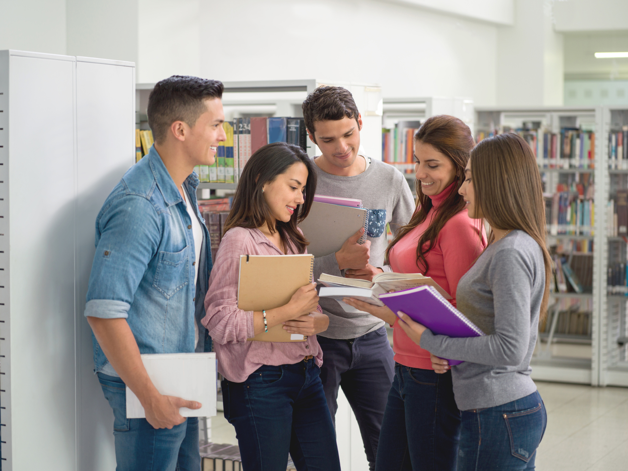 A group of five young adults standing in a library, holding books and notebooks, and engaging in discussion or study together.