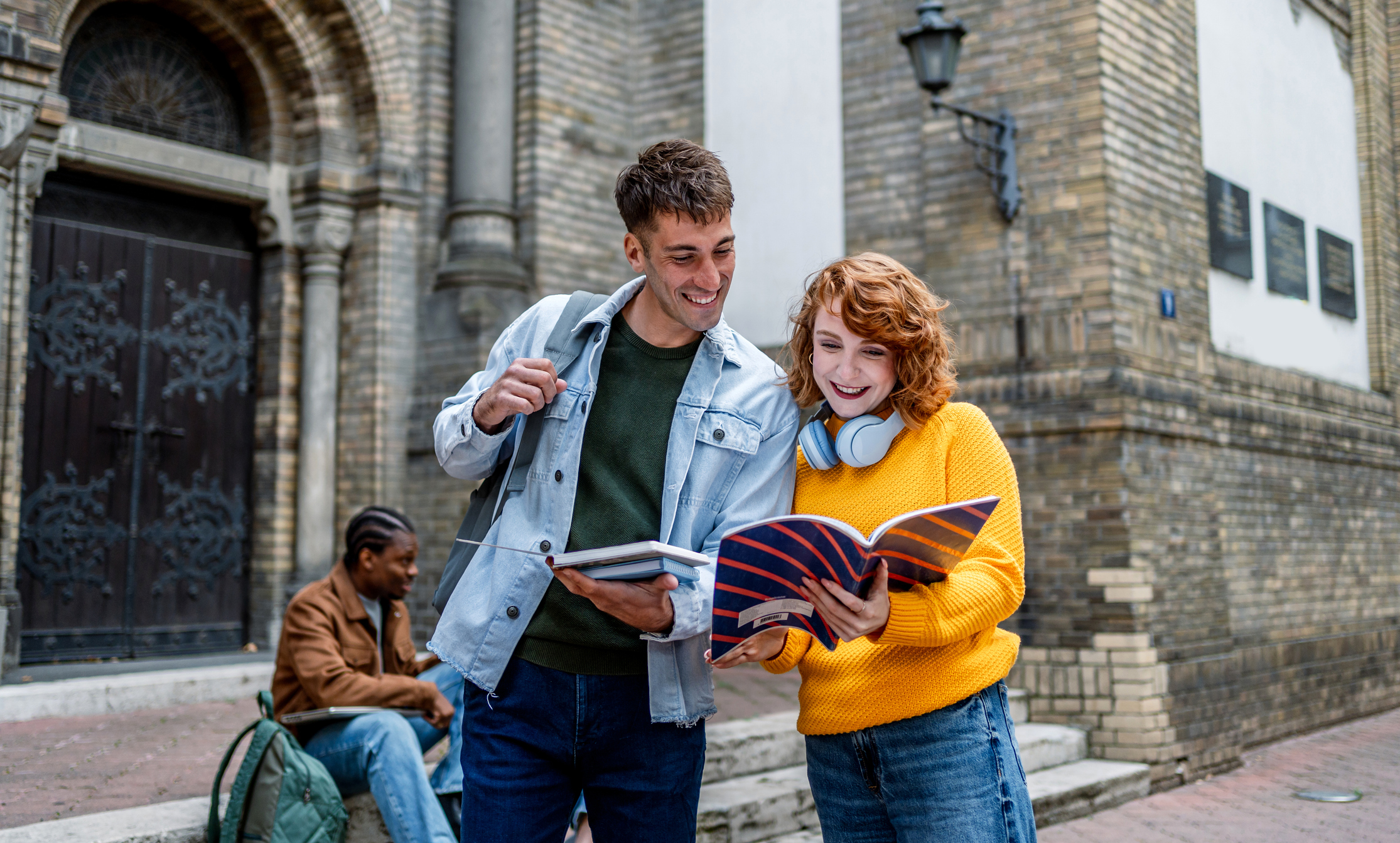 Two students, one wearing a light denim jacket and the other in a yellow sweater with headphones around her neck, smile while looking at notebooks outside an old brick building. Another student sits on steps in the background, engaged with a laptop.