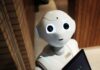 The importance of relationships in times of AI A humanoid robot with large, expressive eyes and a small smile stands in a room with wood-paneled walls, holding a tablet.