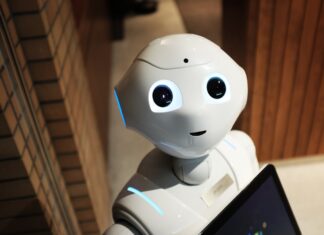 The importance of relationships in times of AI A humanoid robot with large, expressive eyes and a small smile stands in a room with wood-paneled walls, holding a tablet.