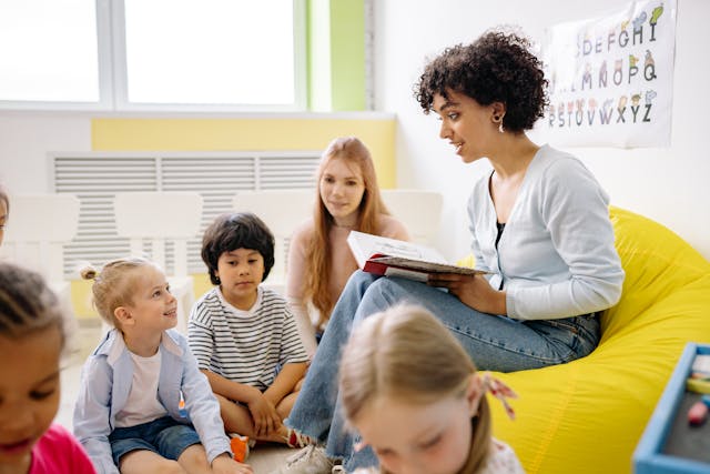 A woman reads a book to a group of young children sitting on a yellow beanbag in a classroom, with an alphabet poster on the wall.