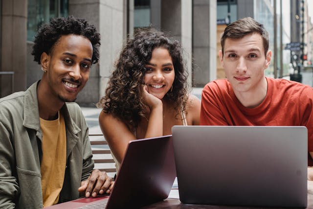 Three people sitting outside at a table with two laptops, smiling at the camera, with urban buildings in the background.