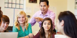 A teacher stands in a classroom surrounded by a group of diverse students, all seated and looking attentively at the teacher, who is gesturing during a discussion.