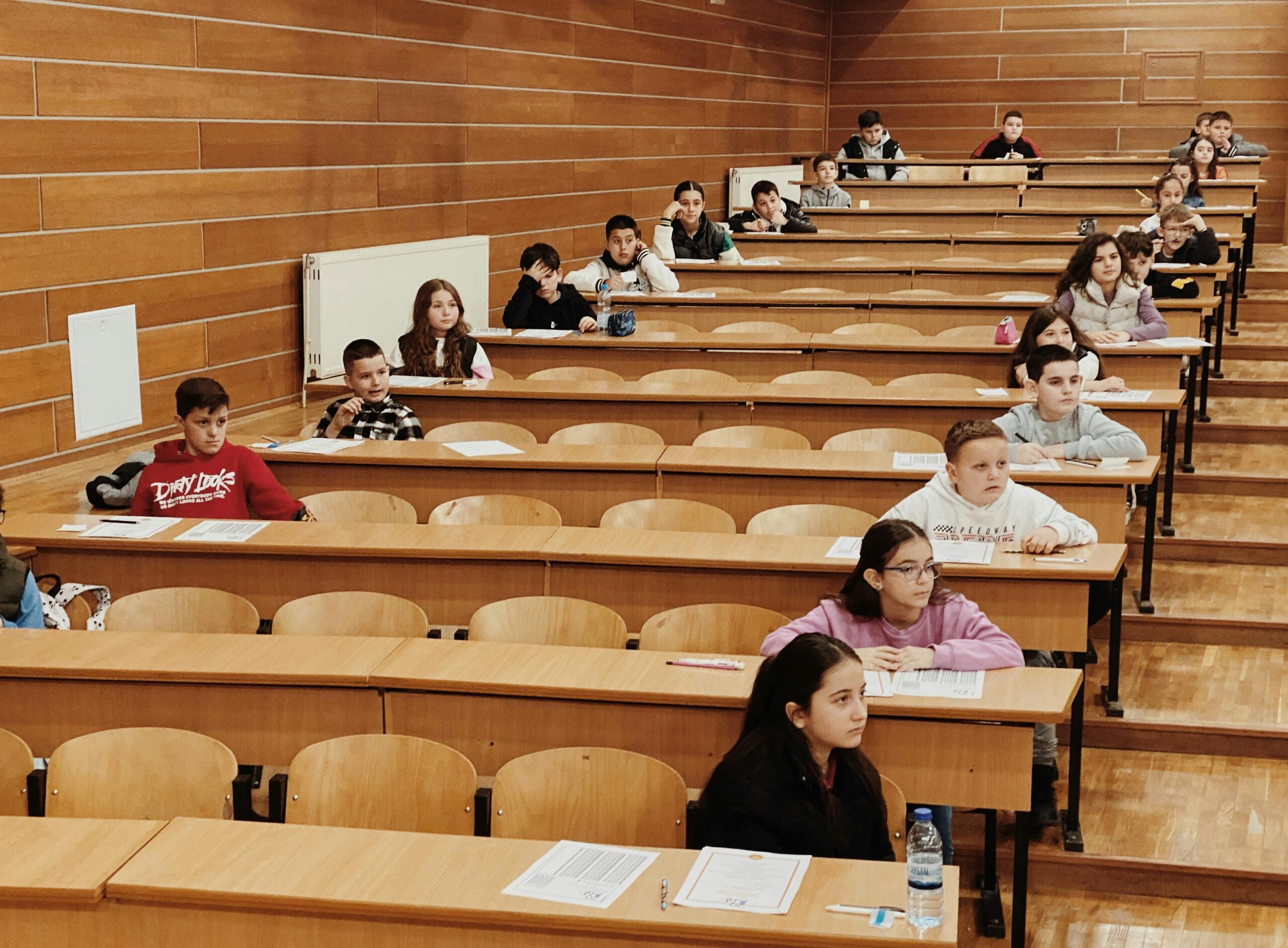 A classroom with students seated at desks arranged in tiered rows, focused on completing papers in front of them. The room has wooden walls and flooring, and some desks have water bottles and pens placed on them.