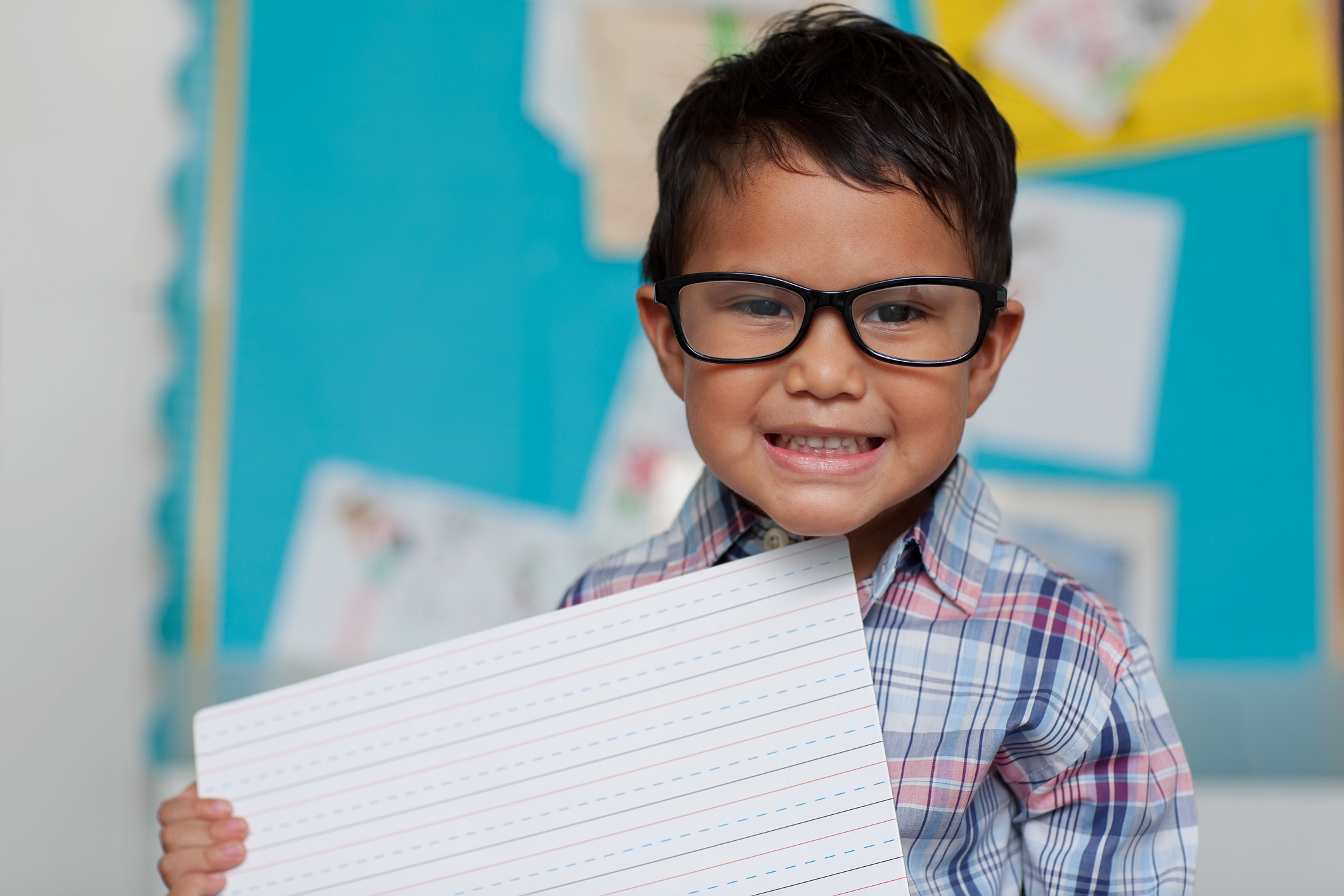 A young boy wearing glasses and a plaid shirt holds up a sheet of lined paper, smiling brightly in a classroom setting with a blue bulletin board in the background.