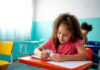Young Learner Exams – Who is it for? A young girl with curly hair in a pink T-shirt writing in a notebook at a classroom desk, with another child in the background.