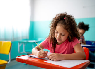 Young Learner Exams – Who is it for? A young girl with curly hair in a pink T-shirt writing in a notebook at a classroom desk, with another child in the background.