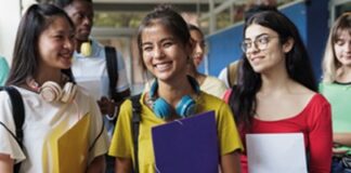 A group of diverse students walking in a school hallway, smiling and holding books and folders.