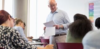 A man in a classroom handing out papers to seated students, with some students writing and others looking at the instructor.