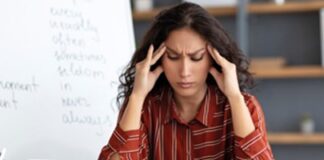 A woman in a striped shirt sits at a desk, holding her temples with a pained expression, with a whiteboard covered in text in the background.